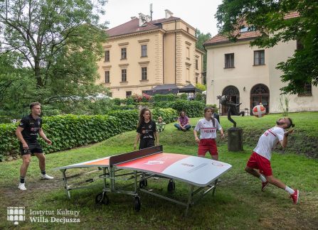 fotografia. dni wyszehradzkie w willi decjusza. mecz teqball na wolnym powietrzu. czworo zawodników wokół stołu ustawionego na trawie. w tle budynek willi decjusza.